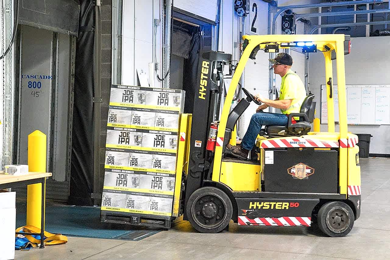 brand-02 A worker drives a Hyster forklift moving Joe IPA boxes in a warehouse.